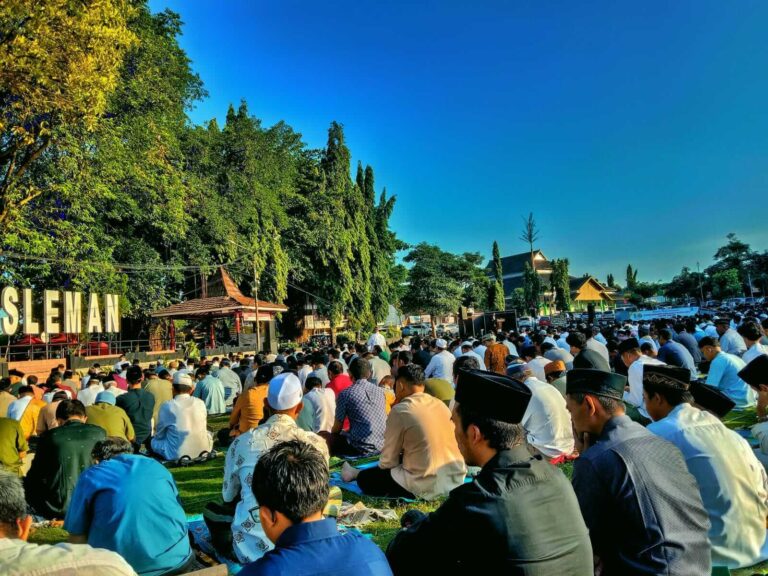 Large crowd seated on prayer mats for an outdoor communal prayer in Sleman, with trees and a stage in the background
